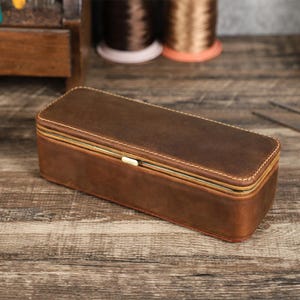 May include: A rectangular brown leather case with a hinged lid. The case is stitched with a light-colored thread and has a metal clasp. The leather has a distressed appearance. The case is likely for storing items.