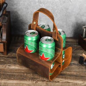 May include: A brown leather beer caddy with a handle, holding four green and silver beer cans. The caddy has individual slots for each can and is made of distressed leather. The cans have a red star logo.
