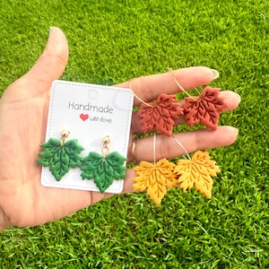 May include: Handmade leaf earrings in green, rust, and yellow. The earrings are in the shape of maple leaves. One pair is on a card that says "Handmade with Love". The earrings are on a background of green grass.