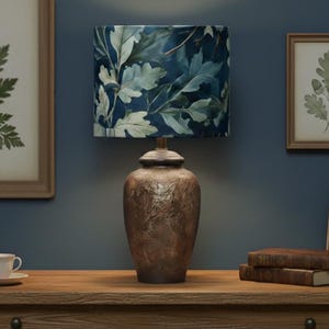 May include: A decorative lamp with a blue and white leaf-patterned lampshade sits on a wooden table. The lamp has a brown, textured base. Two framed botanical prints and a small teacup and saucer are also on the table.