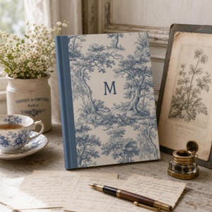 May include: A hardcover journal featuring a blue and white toile de jouy pattern of trees and small animals, with a prominent capital letter M centered on the front cover. The spine is a solid blue color, and the book has a rectangular shape.