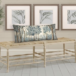 May include: A decorative bench with a woven seat and light wood frame. A long, rectangular pillow with a forest scene in shades of blue and gray rests on the bench. Three framed botanical prints hang above the bench, and a potted olive tree is to the left.