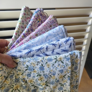 May include: A selection of floral fabric squares in a range of colours, including white, pink, blue, and yellow. The fabrics display diverse floral and dragonfly patterns. The squares are held together, highlighting the variety of designs.