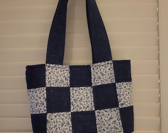 May include: A navy blue and white patchwork tote bag with floral print squares. The bag has long navy blue handles and is hanging against a white shutter background. The tote bag is a handmade item.