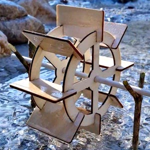 Puede incluir: Una noria de madera clara se encuentra en un arroyo. La rueda, con sus palas, está sostenida por postes de madera. El agua clara refleja la luz del sol. El diámetro de la rueda es de aproximadamente 20 cm.