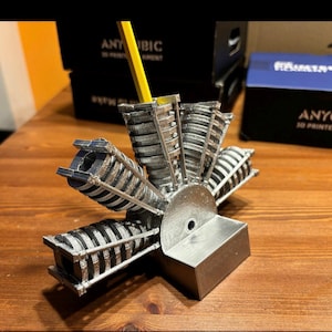 Könnte beinhalten: Ein silberner und schwarzer 3D-gedruckter Stifthalter in Form eines Motors. Ein gelber Bleistift ist in den Halter eingesetzt. Der Stifthalter steht auf einer Holzoberfläche, im Hintergrund befinden sich Anycubic 3D-Druckerfilament-Boxen.