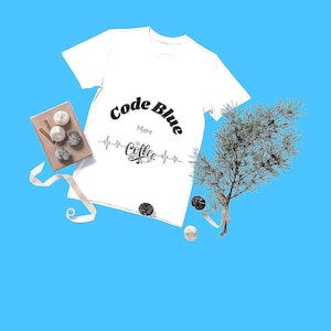 May include: A white t-shirt featuring the text "Code Blue More Coffee" in black. The image includes decorative items like a small gift box, ribbon, and greenery, arranged on a blue backdrop.