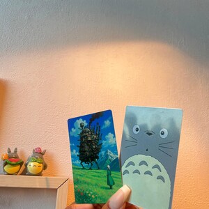 Peut inclure: Deux cartes rectangulaires représentant des personnages d'anime. L'une représente une scène avec un château ambulant, une personne en robe verte et un arrière-plan montagneux. L'autre montre un personnage Totoro gris avec de grands yeux et un ventre blanc.