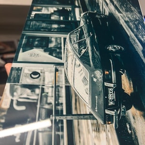 Könnte beinhalten: Schwarz-Weiß-Druck eines Oldtimers, der von vorne und von der Seite gezeigt wird. Das Nummernschild des Autos lautet "E3322 H". Der Hintergrund zeigt eine Stadtansicht mit Gebäuden und Reflexionen.