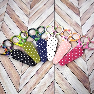 May include: A collection of colorful embroidery scissors with patterned handles and matching protective covers. The scissors feature various designs, including floral, polka dots, and abstract patterns. The handles are in shades of green, pink, blue, and black.