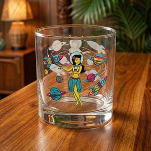May include: A clear glass tumbler featuring a retro-style illustration of a woman in a hula skirt and space helmet surrounded by planets and rockets. The design incorporates bright colors like blue, pink, yellow, and green.