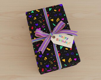 May include: A rectangular gift box wrapped in black paper featuring a colorful pattern of small hearts, triangles, and geometric shapes. A multi-colored striped ribbon is tied in a bow around the box, holding a white tag that reads Happy Birthday.