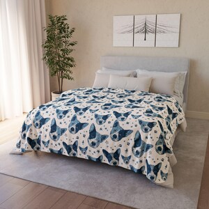 May include: A cream-coloured blanket featuring a repeating pattern of blue dog faces, paw prints, and stars. The blanket is on a bed with white pillows and a grey headboard. The room has a neutral colour scheme.