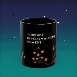 Puede incluir: Taza de cerámica negra con el texto "It's not DNS, There's no way it's DNS, It was DNS." Diseño de rama de flor de cerezo rosa. La taza es cilíndrica y tiene un acabado brillante.