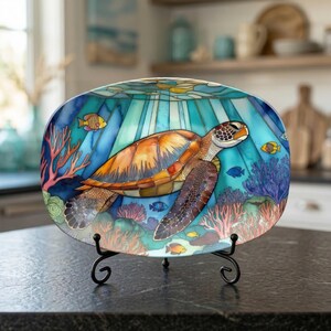 May include: An oval decorative plate featuring a sea turtle swimming underwater. The plate has a colorful design with coral, fish, and light rays. The plate is displayed on a black metal stand.