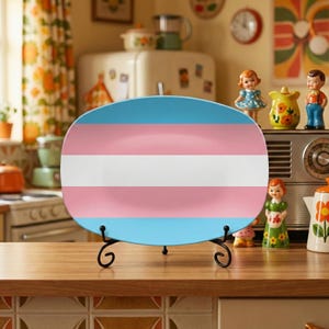 May include: An oval ceramic plate with a blue, pink, and white horizontal striped design, displayed on a black metal stand. The plate is set on a wooden countertop in a kitchen setting, with vintage decor in the background.