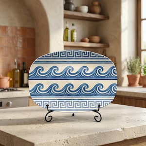 May include: An oval decorative plate with a blue wave pattern and Greek key border, displayed on a black metal stand. The plate has a cream-colored background. The plate is on a kitchen countertop.