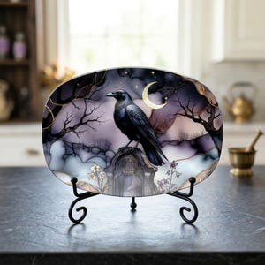 May include: An oval decorative plate featuring a black raven perched on a tombstone, set against a night sky with a crescent moon and stars. The plate has a dark, gothic theme with gold accents and is displayed on a black metal stand.