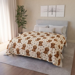 Puede incluir: Una manta color crema con un patrón repetido de ilustraciones de caniches marrones, huellas de patas y pequeños corazones. La manta está sobre una cama con almohadas blancas y un cabecero gris. La habitación tiene tonos neutros.