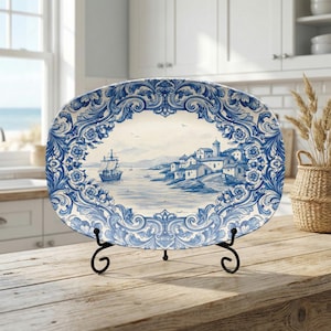 May include: Oval ceramic serving platter with a blue and white coastal scene design. The platter features a detailed illustration of a seaside village and a sailing ship, framed by ornate blue floral patterns. It is displayed on a black metal stand.