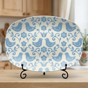 May include: Oval serving platter with a cream background and a repeating pattern of blue birds, flowers, and vines. The platter is displayed on a black metal stand, suitable for serving food or as a decorative piece.