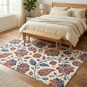 May include: A rectangular area rug with a cream background and a floral pattern in shades of red and blue. The rug is placed in a bedroom setting, with a wooden bed frame and bench visible.