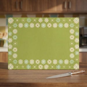 Può includere: Un tagliere rettangolare verde con un bordo di fiori di margherita bianchi, azzurri e color crema. Un coltello in acciaio inossidabile con manico in legno è appoggiato sulla superficie di legno davanti al tagliere.