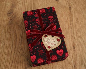May include: A wrapped gift box with a black background featuring red roses, skulls, and hearts. A dark red velvet ribbon and bow secure the box, with a heart-shaped tag that reads "To My Valentine".