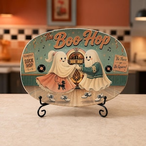May include: An oval decorative plate on a black metal stand featuring two ghosts dancing. The text reads The Boo Hop, Tonite! Sock Hop 7 to 11, and Be There or Be Square! The ghosts wear a pink poodle skirt and a letterman jacket, with a jukebox.