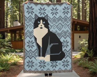 May include: A woven throw blanket featuring a black and white cat design on a light blue background with gray snowflake and geometric patterns. The blanket has a fringed edge and is held up in front of a house and trees.