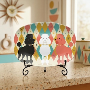 Peut inclure: Une assiette décorative ovale avec trois illustrations de caniches noirs, blancs et rose corail. L'assiette présente un motif géométrique en losanges colorés et est présentée sur un support en métal noir. C'est un objet décoratif.