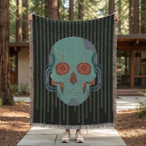 May include: A woven throw blanket featuring a teal skull design with red eyes and circuit board details. The skull is set against a black background with vertical green lines. The blanket has fringed edges.