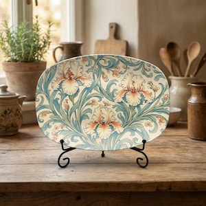 May include: An oval decorative plate with a floral iris design in cream, blue, and peach tones. The plate is displayed on a black metal stand. The background includes a wooden table, a window, and kitchen utensils.