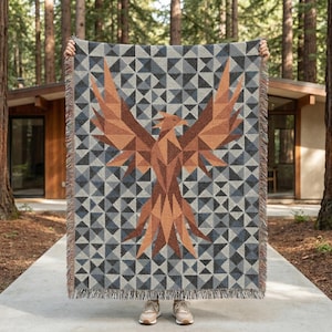 May include: A woven blanket featuring a geometric design with a large phoenix in shades of brown and orange. The background is a pattern of gray and blue triangles. The blanket has a fringed edge and is held up in front of a house and trees.
