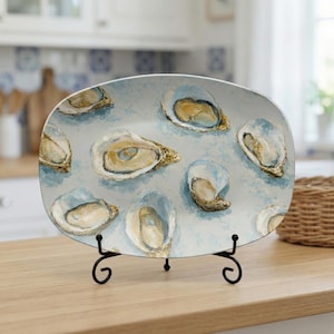 May include: An oval ceramic serving platter with a light blue background and painted oyster illustrations. The oysters are depicted with white, gold, and blue hues. The platter is displayed on a black metal stand.