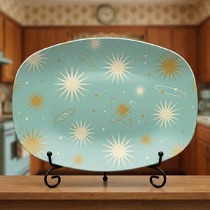 May include: A light blue, oval-shaped serving platter with a retro space-themed design. The platter features starbursts, planets, and atomic shapes in cream and gold. It is displayed on a black metal stand.