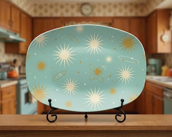 May include: A light blue, oval-shaped serving platter with a retro space-themed design. The platter features starbursts, planets, and atomic shapes in cream and gold. It is displayed on a black metal stand.