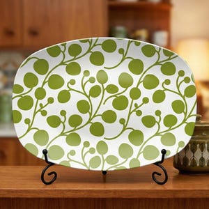 May include: White oval serving platter with a green leaf pattern. The platter is displayed on a black metal stand. The design features a repeating pattern of green leaves and stems, creating a botanical theme.