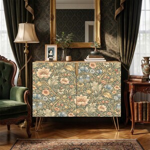May include: A wooden cabinet with a floral pattern in shades of blue, peach, and yellow. The cabinet has gold hairpin legs and is set against a dark green wall. A gold-framed mirror hangs above the cabinet.