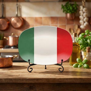 Puede incluir: Un plato ovalado decorativo con los colores de la bandera italiana: verde, blanco y rojo. Se muestra sobre un soporte de metal negro. El fondo es una cocina con ollas de cobre y botellas de aceite de oliva.