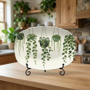 May include: An oval serving platter featuring a botanical design of hanging plants in black pots with trailing green leaves. The platter is cream-colored and displayed on a black metal stand, perfect for serving food or as a decorative piece.