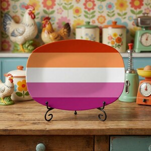 May include: An oval ceramic plate featuring a horizontal striped design in orange, white, pink, and magenta, presented on a black metal stand. The plate is displayed on a wooden surface.