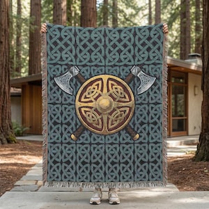 May include: A woven blanket with a teal and black Celtic knot pattern. The center features a gold and brown shield with two crossed axes. The blanket has a fringed edge and is held up in front of a natural background.