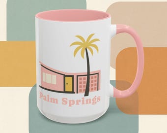 Op de afbeelding: Witte keramische mok met een roze binnenkant en handvat. De mok heeft een roze huis in mid-century modern stijl met een gele palmboomafbeelding en de woorden "Palm Springs" in roze.