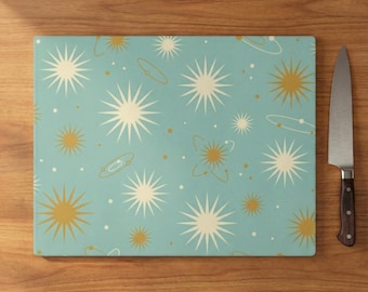 May include: A rectangular cutting board with a retro space-themed design. The board features a light blue background with starbursts and orbital illustrations in cream and gold. A stainless steel knife with a wooden handle rests on the wooden surface.