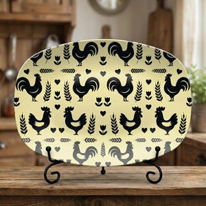 Puede incluir: Una fuente ovalada con fondo amarillo y un patrón repetido de gallos negros. La fuente se exhibe sobre un soporte de metal negro, adecuado para la decoración de cocina o comedor.