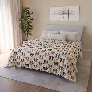 Può includere: Una coperta beige con un motivo ripetuto di facce di cani in vari colori, tra cui marrone, nero e blu. Piccoli cuori neri sono sparsi nel design. La coperta è su un letto con cuscini bianchi.