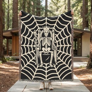 Könnte beinhalten: Schwarze Decke mit cremefarbenem Spinnennetz-Design und einem Skelettkönig auf einem Thron. Das Skelett trägt eine Krone und hält ein Zepter. Die Decke hat einen Fransenrand.