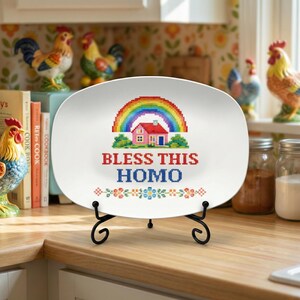May include: White oval decorative plate with a cross-stitch design featuring a rainbow, house, and the text "BLESS THIS HOMO" in red and blue. The plate is displayed on a black metal stand.