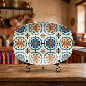 May include: An oval serving platter with a floral pattern in shades of blue, orange, and cream. The platter is displayed on a black metal stand, showcasing its decorative design. The kitchen setting adds a rustic charm.